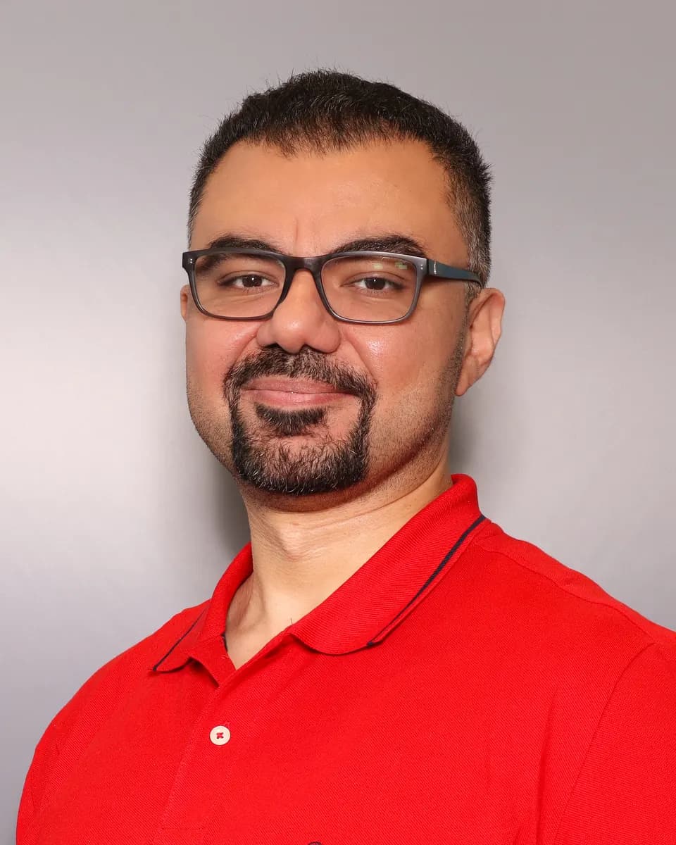 Portrait of Anas Aboreeda, smiling, wearing glasses and a red polo shirt against a grey backdrop